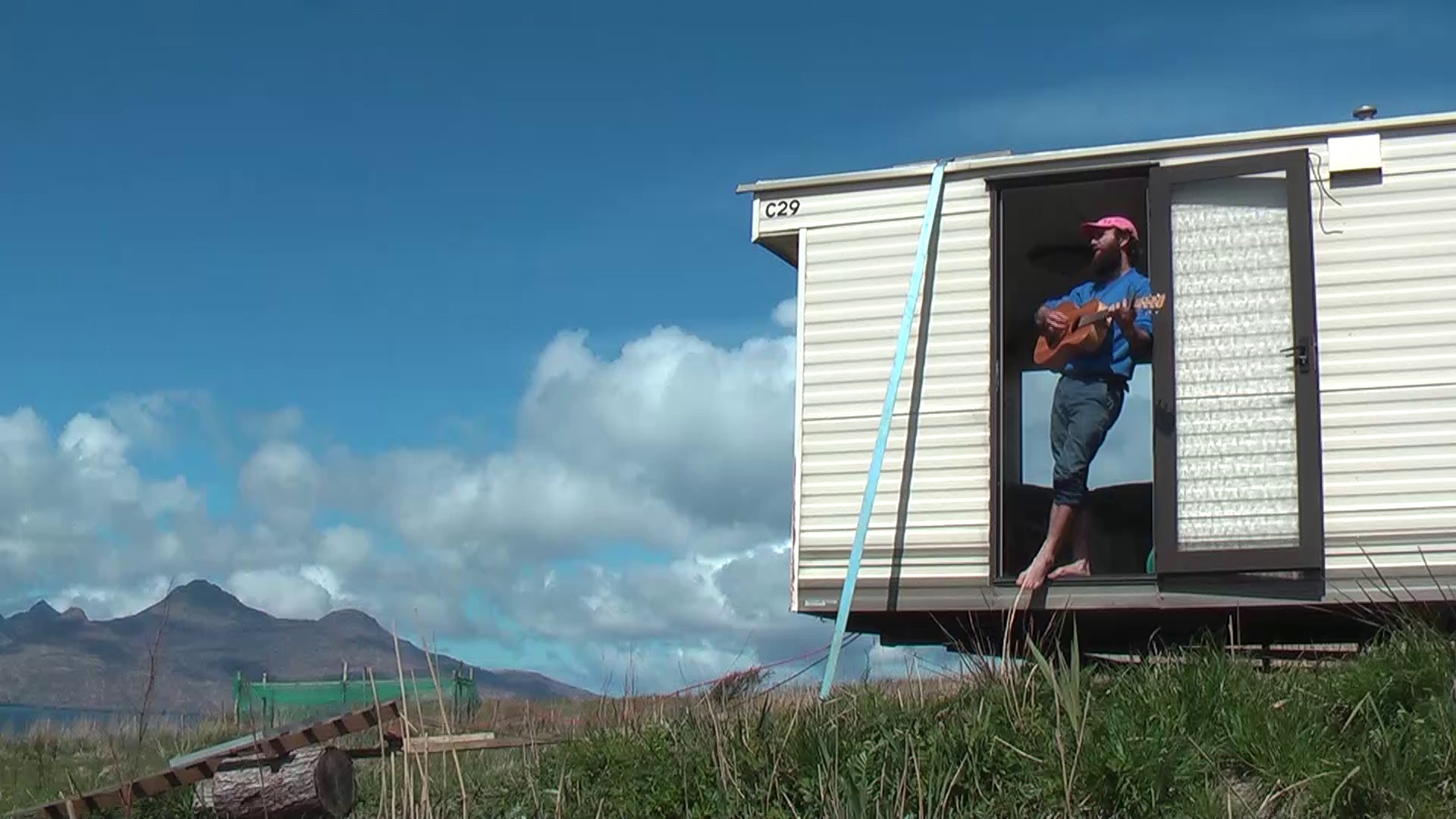 A photograph of Ed Dickinson playing guitar, standing in the open door of a static caravan. The caravan is in a field, on a clear day with the rugged peaks of the Isle of Rum in the background.
