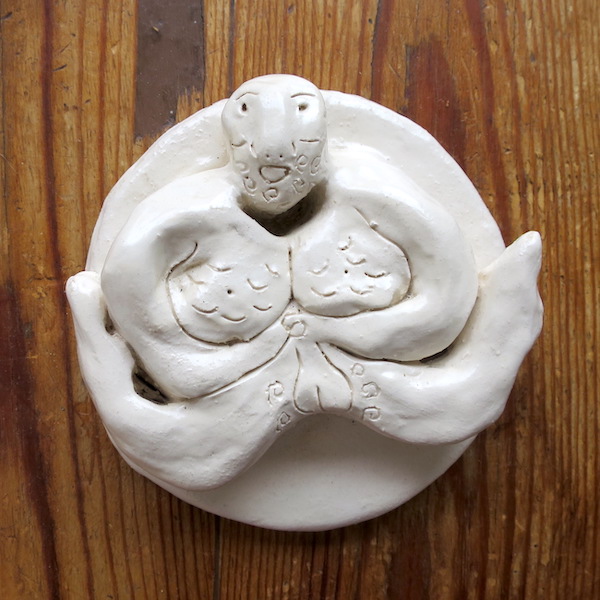 A photo of a ceramic figure on a wooden surface. The figure is man with his legs spread either side of him, grabbing his penis with both hands.